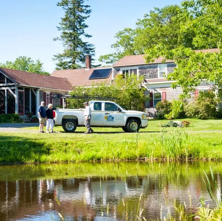 A Mid-Coast Energy Systems technician speaks with two homeowners beside a branded white pickup truck in front of a shingled house in Midcoast Maine. The home features heat pump and solar installations, surrounded by trees and reflected in a nearby pond.